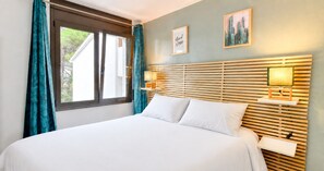A hotel room with a bed, a window with curtains, a headboard with a light, and two wall art pieces. - Apartment with pool, AC and tennis · Calella (Calella de Palafrugell)