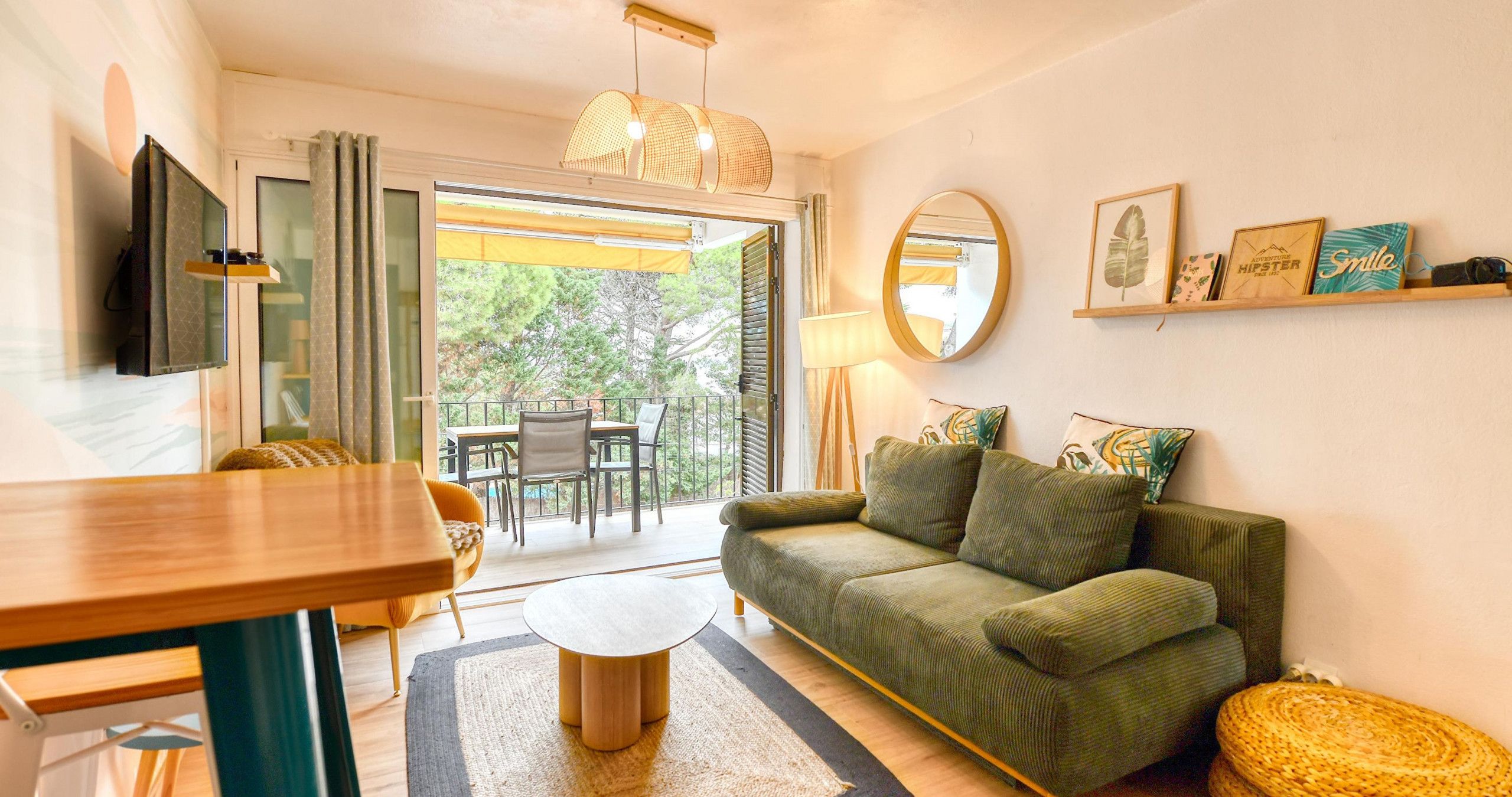 A living room with a sofa, a round coffee table, a TV mounted on the wall, and a balcony with a table and chairs.
