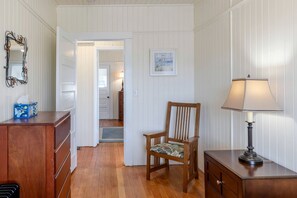 A room with a wooden dresser, a chair with a floral cushion, a lamp, a framed picture on the wall, and a door leading to another room. - Classic Beach House with Game Room & Firepit Near Seaside Beach in Seaside, Oregon (Seaside)