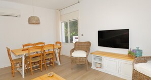 Ein Essbereich mit einem Holztisch und Stühlen, einem Fernseher an der Wand und einem Fenster mit Blick auf Grünflächen.