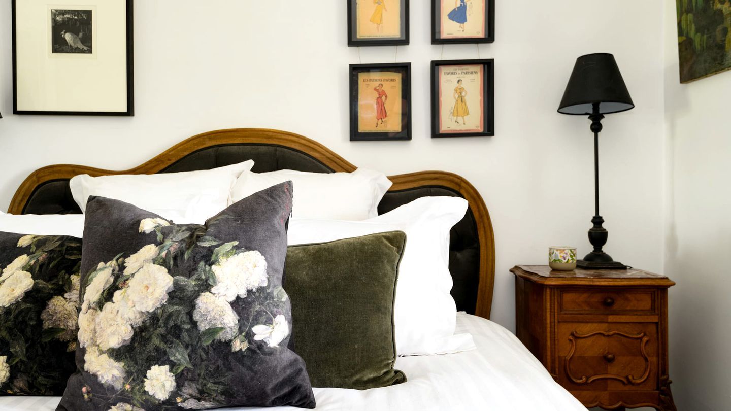 Un lit bien fait, agrémenté d’oreillers à motifs floraux, une tête de lit en bois, une table de chevet avec une lampe et des tableaux encadrés accrochés au mur.