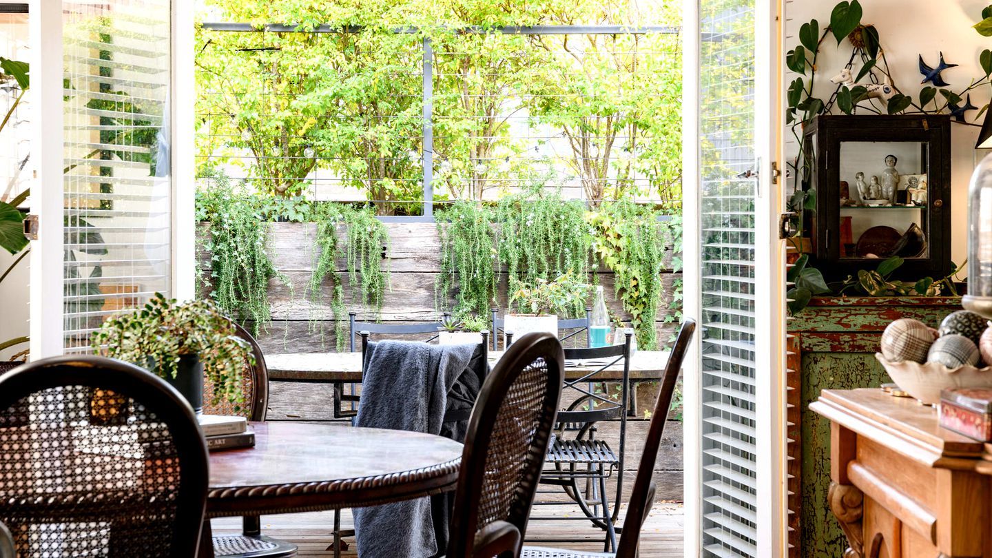 An outdoor patio with a round table and chairs, a wooden fence, and a view of a garden with trees and plants.