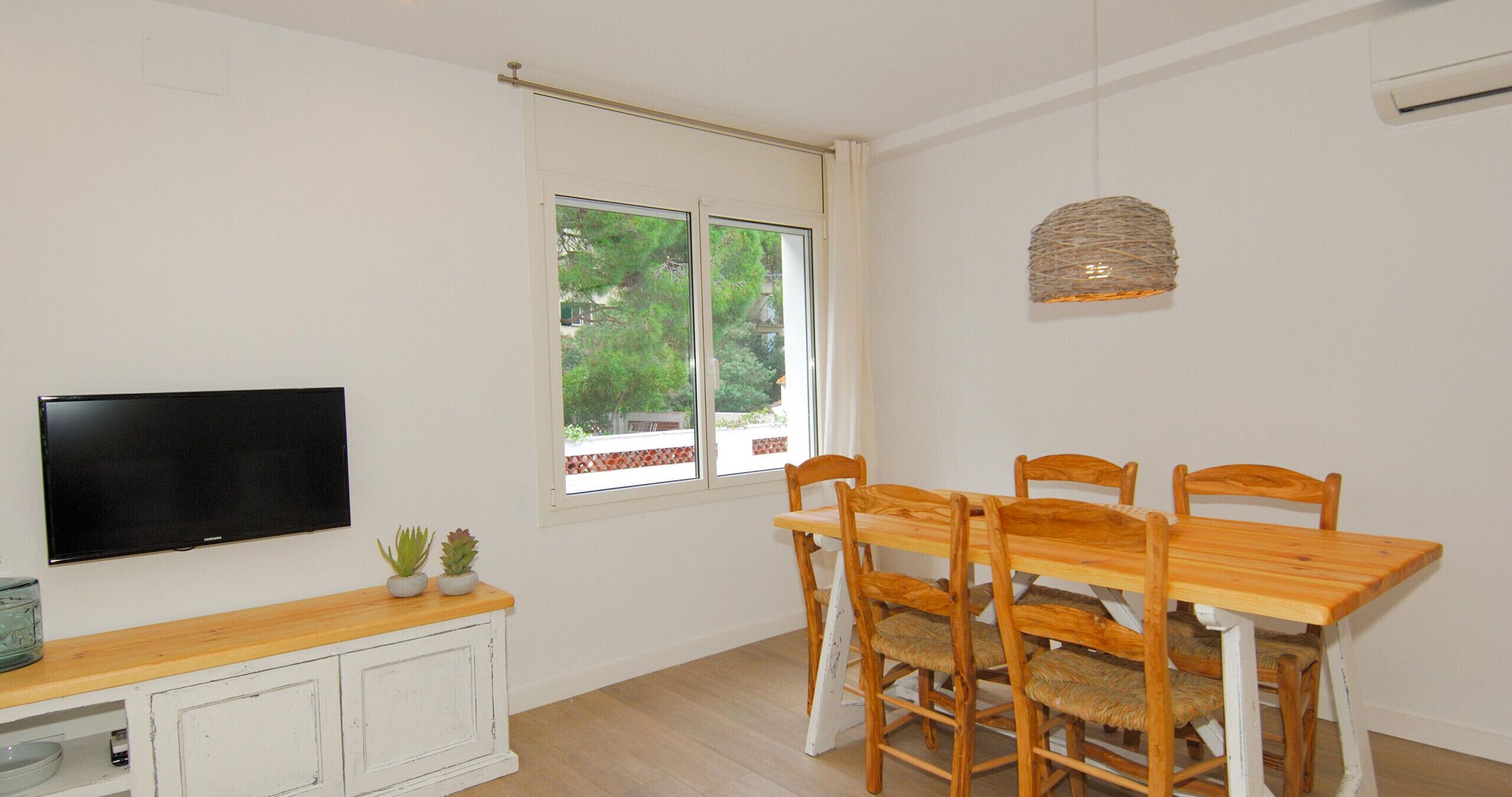 A dining area with a wooden table and chairs, a television on the wall, and a window with a view of trees.