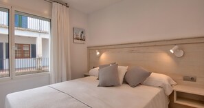 Ein Schlafzimmer mit Bett, Kissen, Nachttisch, Wandleuchte, Vorhängen am Fenster und Blick auf einen Balkon.