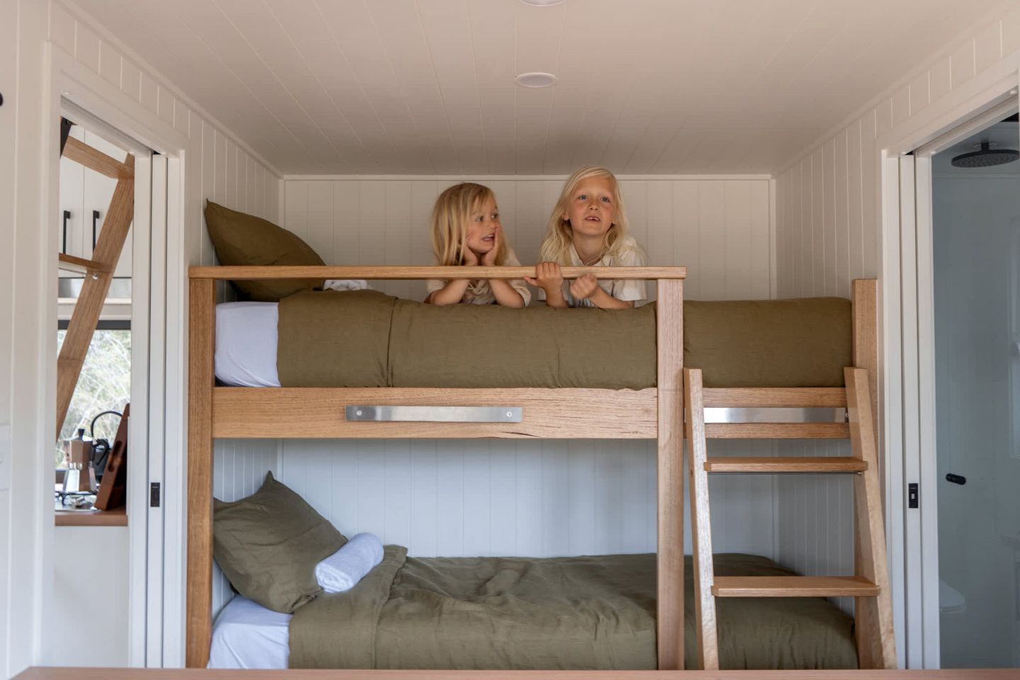 A bunk bed room with two children sitting on top, wooden railings, and white walls.