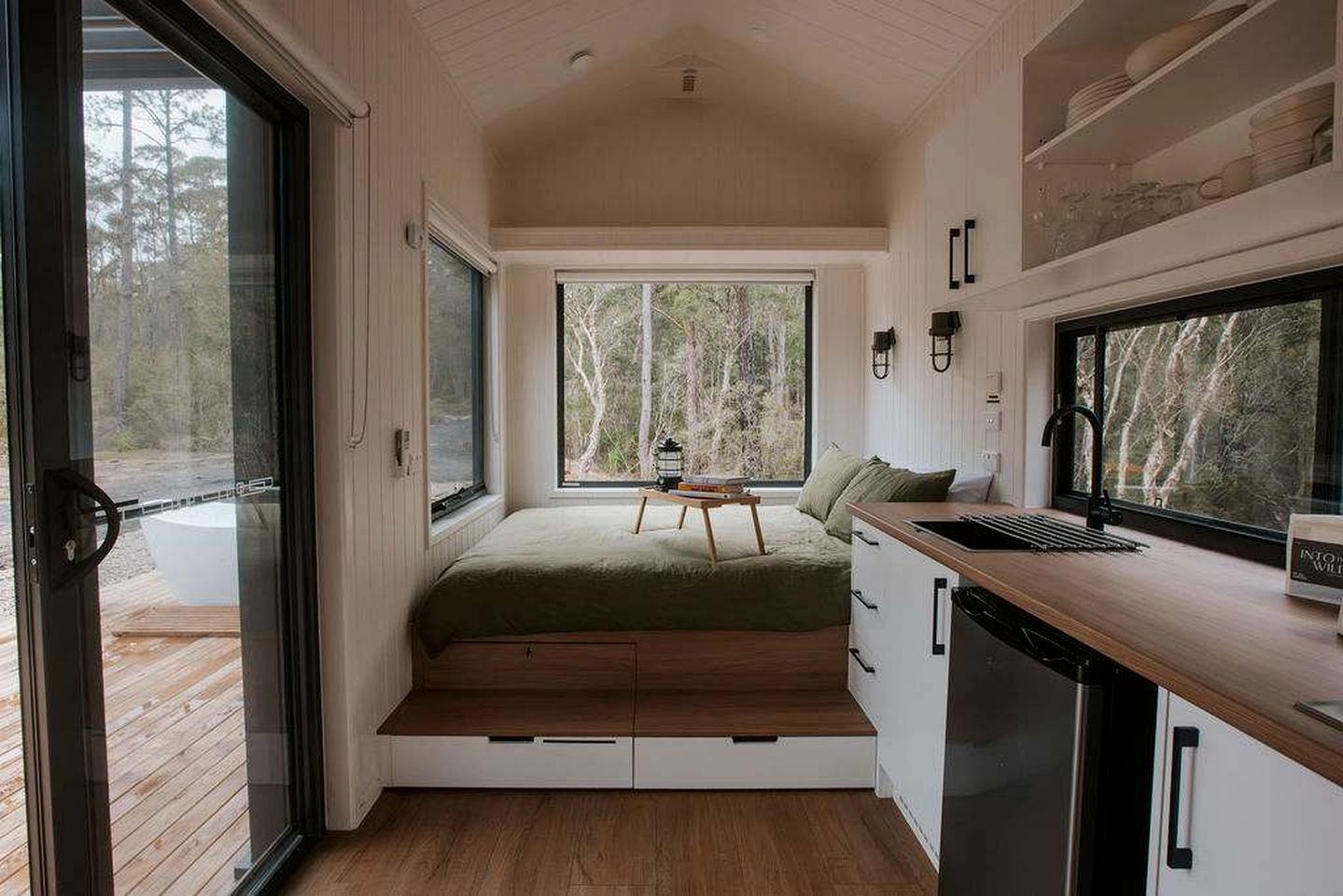 A compact room with a bed, a small table, a sink, and a window overlooking a forest.