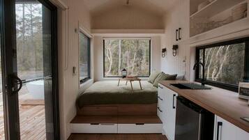 A compact room with a bed, a small table, a sink, and a window overlooking a forest.