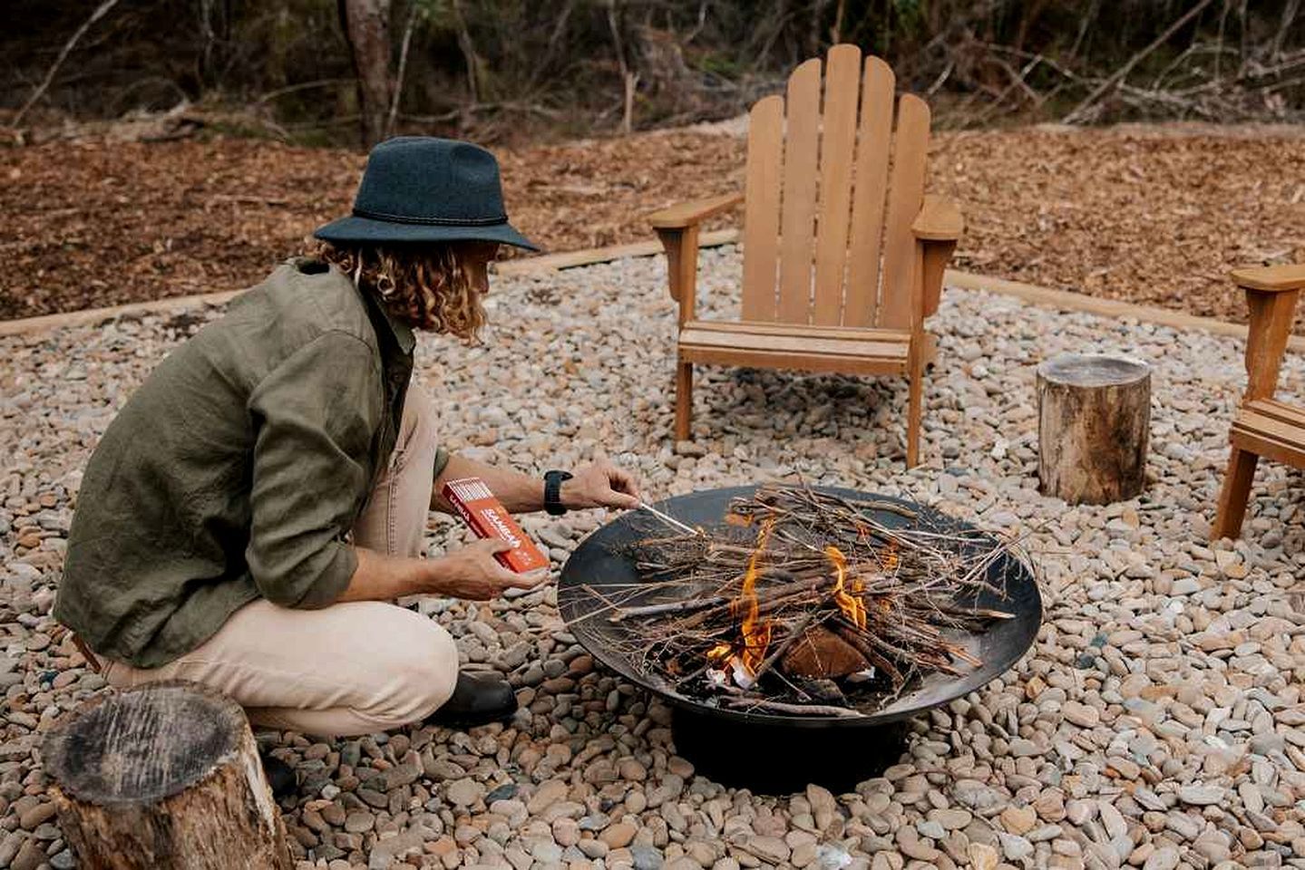A person tending to a campfire in a rocky outdoor setting with Adirondack chairs and a log fire pit.