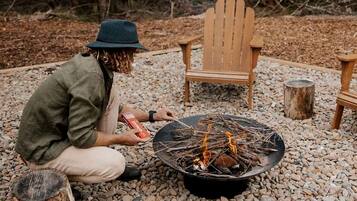 A person tending to a campfire in a rocky outdoor setting with Adirondack chairs and a log fire pit.