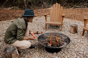 Una persona si occupa di un fuoco da campo in un ambiente naturale roccioso, con poltrone Adirondack e un braciere in legno.