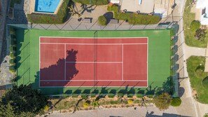 Eine Vogelperspektive auf einen Tennisplatz mit umgebender Grünfläche und einem Schwimmbecken.