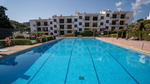 En swimmingpool med kristallklart blått vatten, omgiven av ett välskött område med bänkar och en byggnad i bakgrunden.