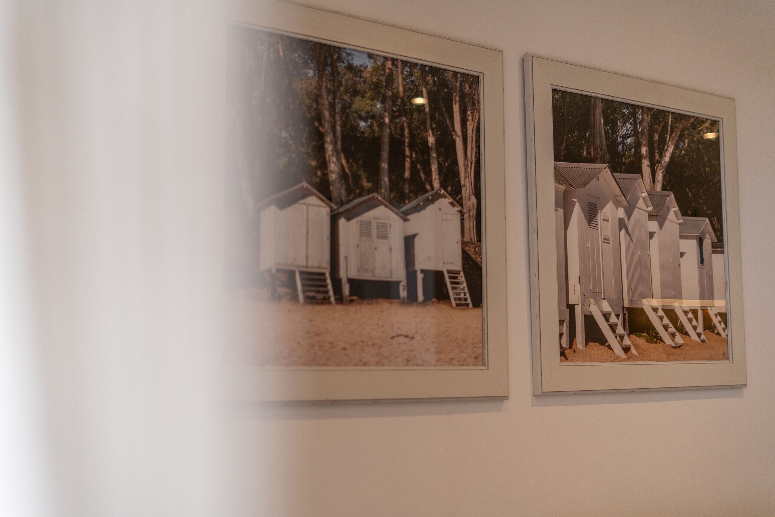Two framed photographs of beach huts with ladders and a sandy ground.