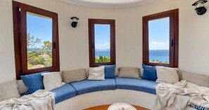 A curved sofa with multiple cushions, three windows with wooden frames, and a view of the sea. - Apartment with Beautiful Sea Views and Pool in Calella · Perfect to Relax (Calella de Palafrugell)