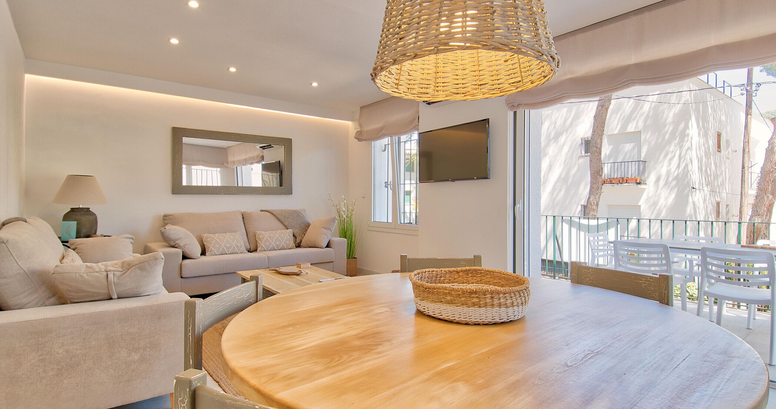 Una sala de estar moderna con una mesa redonda de madera, un sofá, un espejo grande y un balcón con sillas blancas.