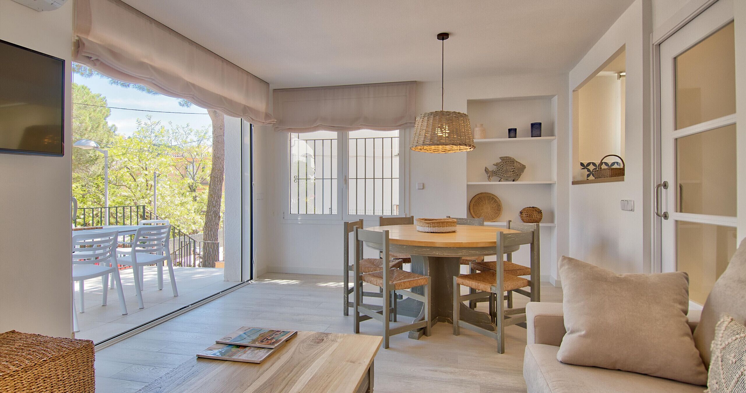 Un comedor con una mesa redonda y sillas, un sofá con cojines, una lámpara colgante y vistas al exterior a través de un ventanal corredero.