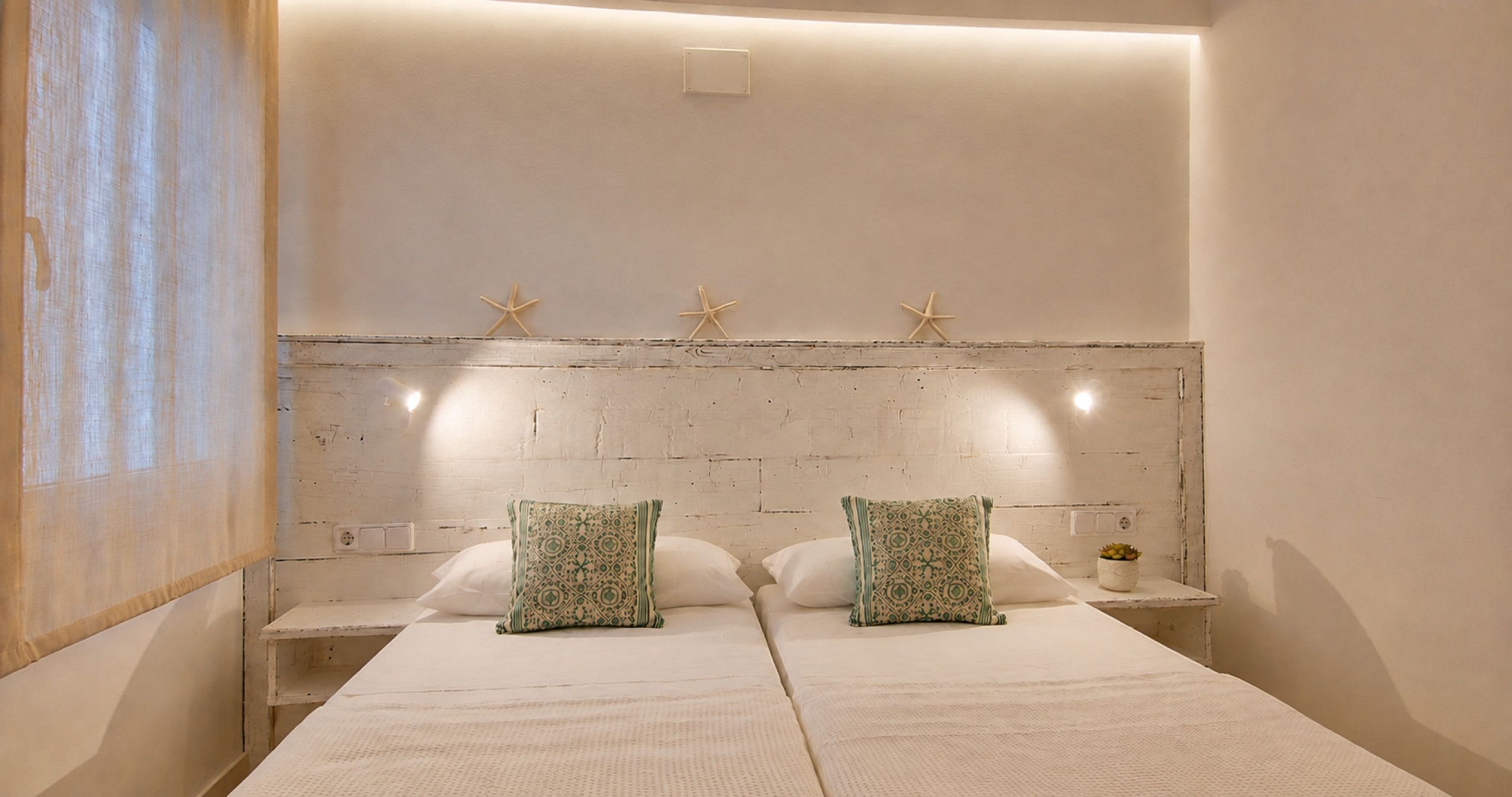 Una cama doble con dos almohadones decorativos, una mesita de noche con una planta pequeña y una pared adornada con tres estrellas de mar.