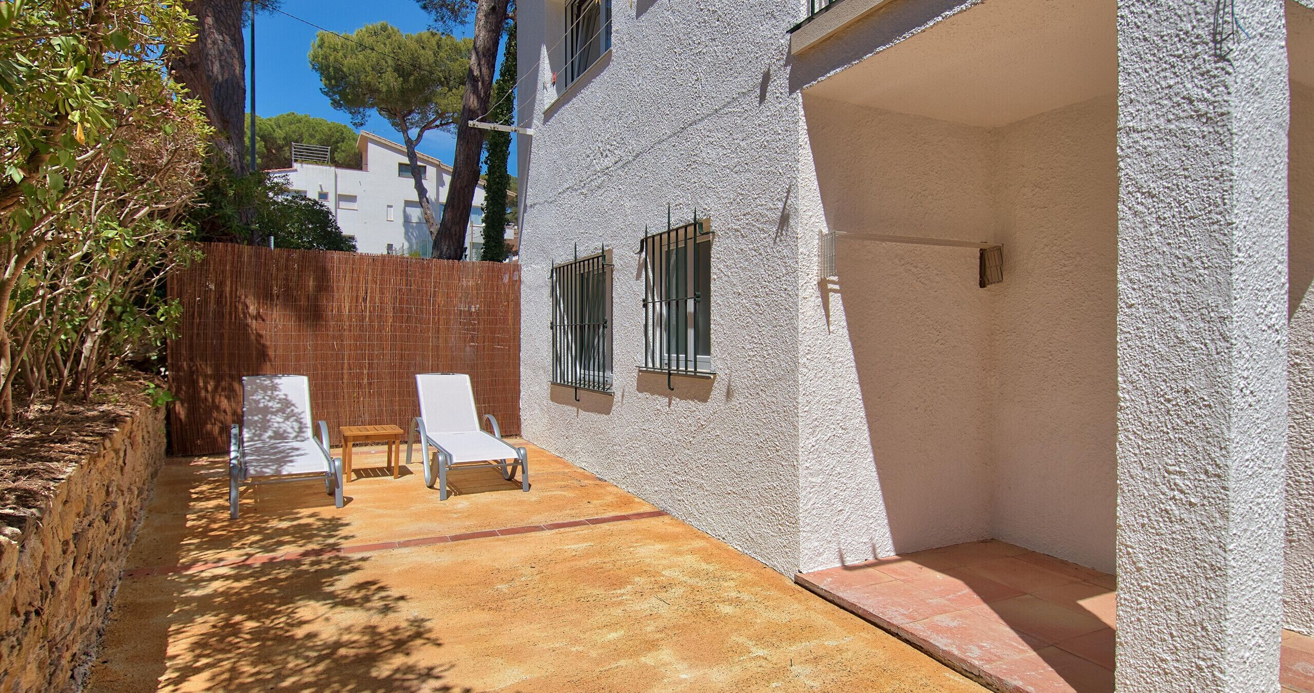 A sunlit outdoor area with a white wall, two lounge chairs, a small wooden table, and a metal railing.
