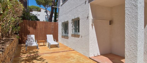 Un espacio exterior iluminado por el sol con una pared blanca, dos sillones de descanso, una pequeña mesa de madera y una barandilla de metal.
