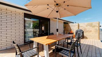 An outdoor patio with a wooden table and chairs, a beige umbrella, and a barbecue grill.