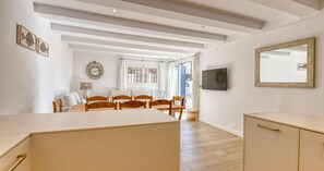 Ein Zimmer mit einem Holztisch, Stühlen, einem an der Wand befestigten Fernseher und einem großen Fenster mit Vorhängen.