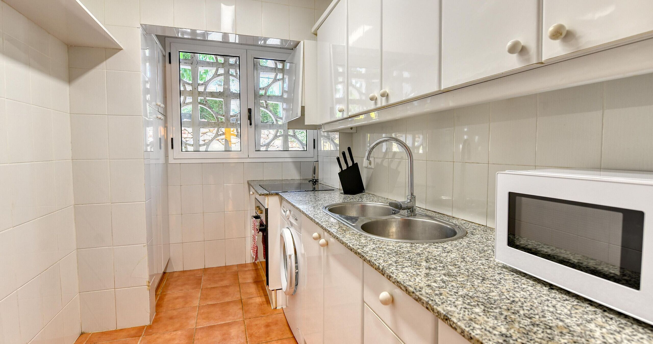 Une cuisine avec des meubles blancs, un évier en acier inoxydable, un four à micro-ondes et une fenêtre donnant sur un paysage verdoyant.