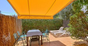 Eine Terrasse im Freien mit Tisch, Stühlen und einer Liege, außerdem ein gelbes Sonnensegel.