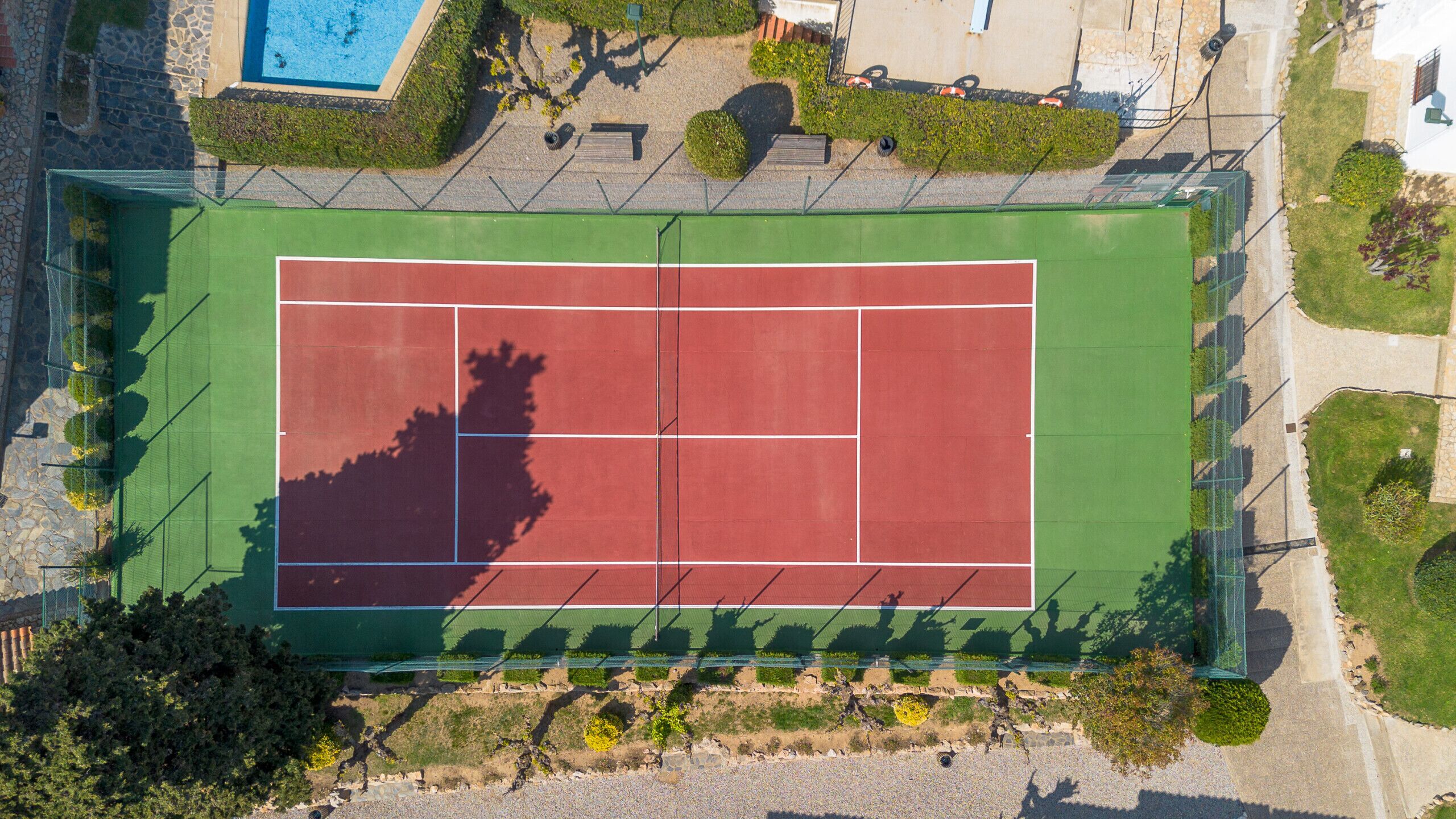 An aerial view of a tennis court with surrounding greenery and a pool.