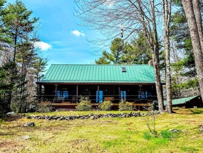 A cabin with a green roof, surrounded by trees, and a stone wall in front. - Waterfront Cabin Retreat with Deck Swing in Lamoine Point, Maine (Lamoine Point)