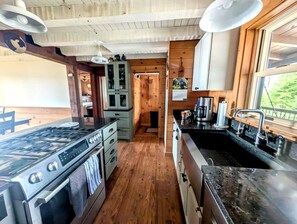 A kitchen with a gas stove, wooden cabinets, and a large window. - Waterfront Cabin Retreat with Deck Swing in Lamoine Point, Maine (Lamoine Point)