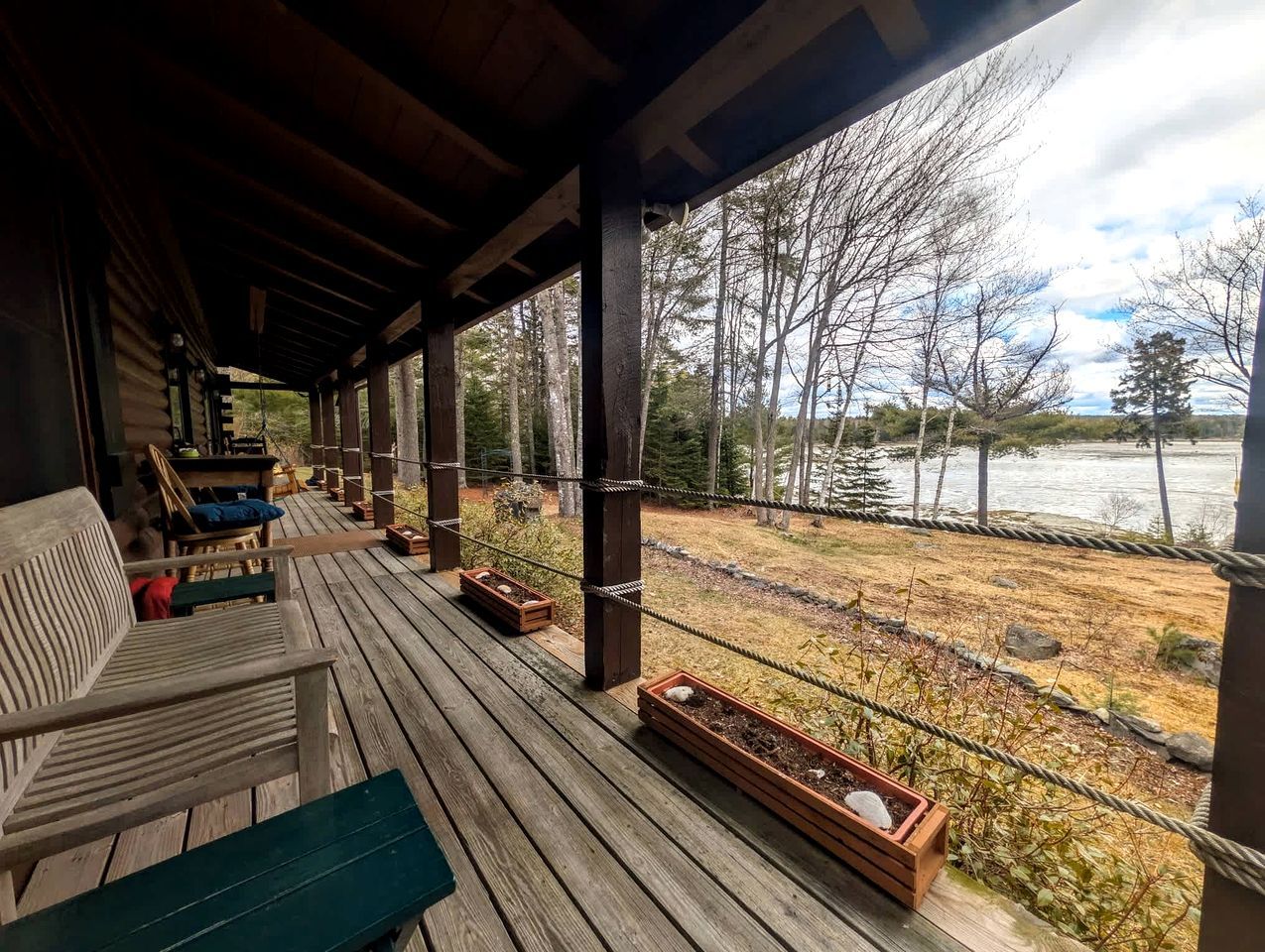 Terraza de madera con mobiliario exterior y macetas, con vista a un río y zona boscosa.