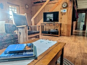 A cozy cabin interior with a wooden staircase, a television, a table with books, and a floral-patterned chair. - Waterfront Cabin Retreat with Deck Swing in Lamoine Point, Maine (Lamoine Point)