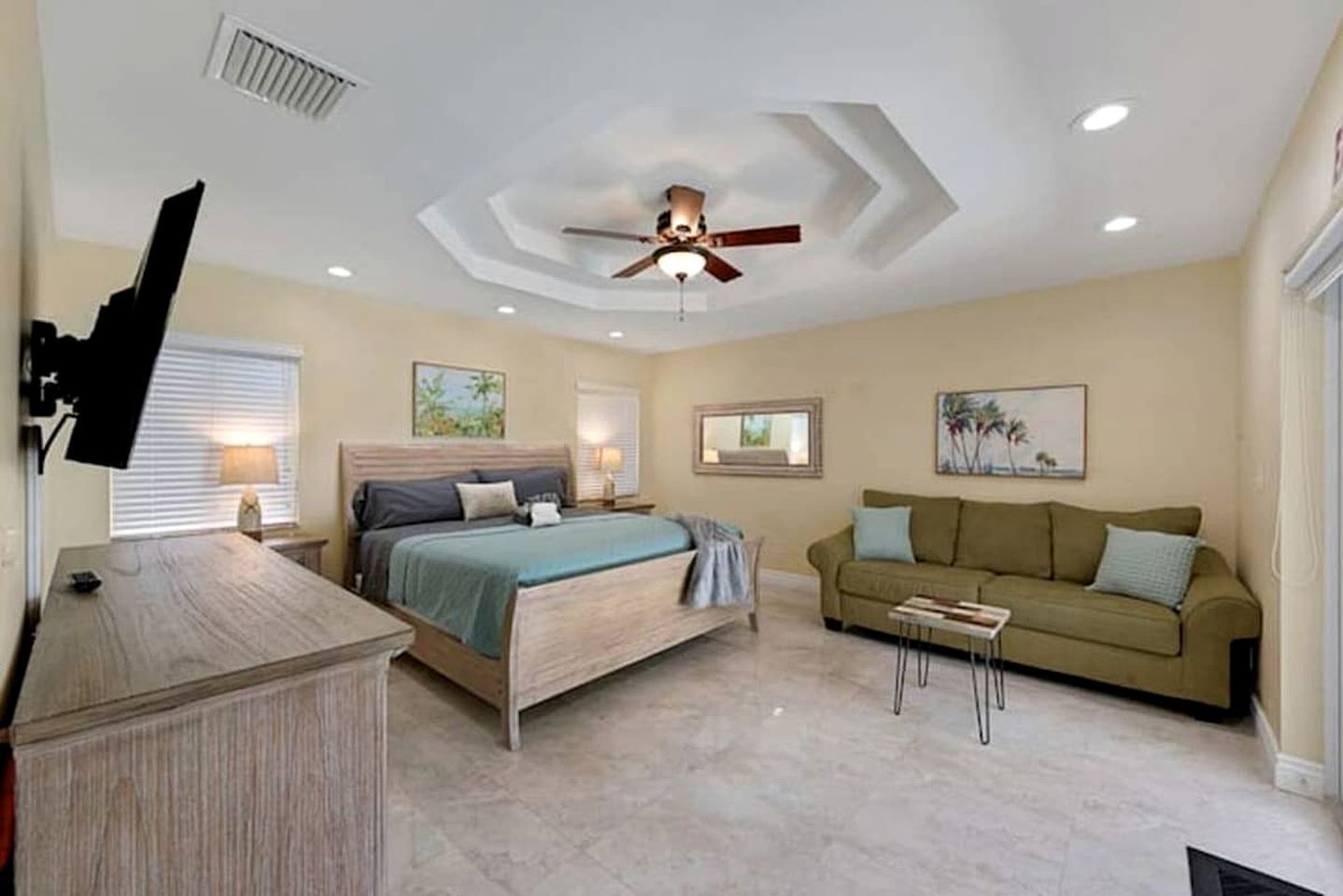 A bedroom with a bed, a ceiling fan, a nightstand, a dresser, a window with blinds, and a wall-mounted TV.