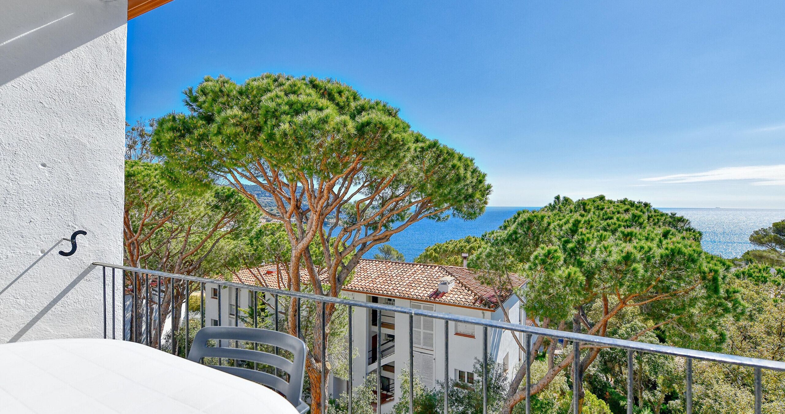Un balcon doté d’une balustrade, donnant sur une zone côtière avec des arbres et un bâtiment dont le toit est recouvert de tuiles.