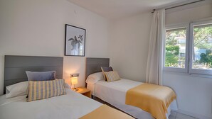 A hotel room with two beds, a nightstand, a lamp, a framed picture on the wall, and a window with curtains. - Sea View Apartment in Calella with Parking Included (Calella de Palafrugell)