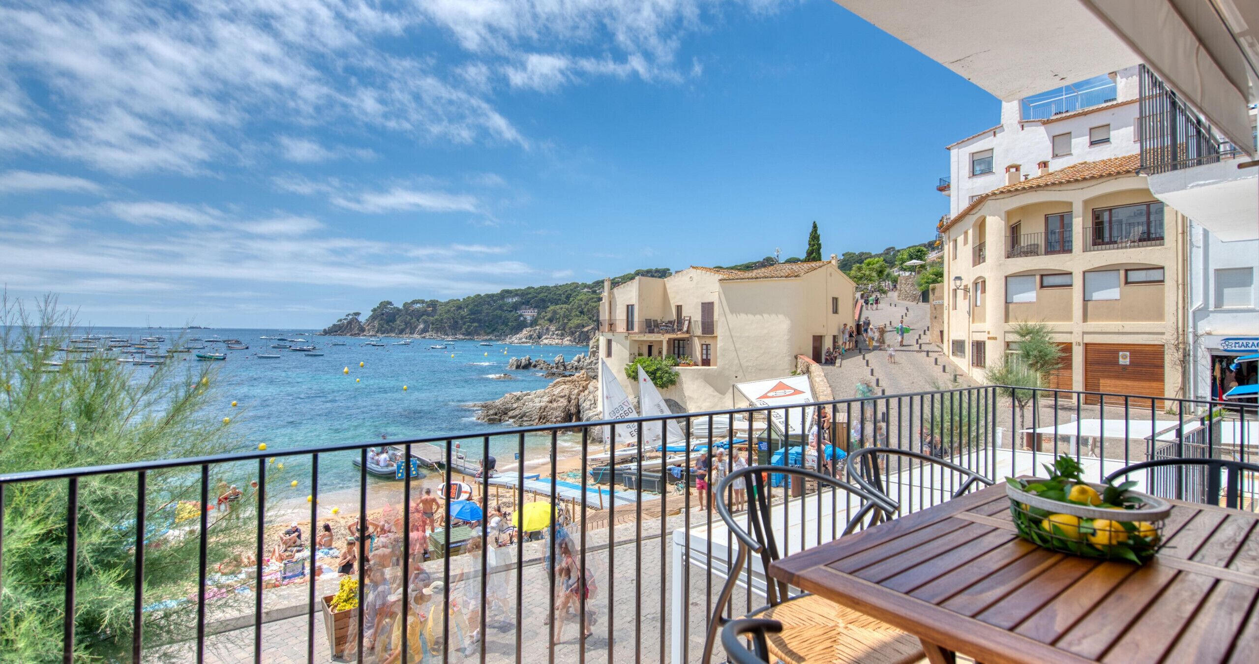 A balcony with a view of a beach, a wooden table with a fruit basket, and a building with a balcony.