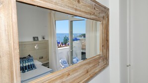 A room with a wooden-framed window, a bed with blue and white bedding, a bedside table, and a view of the sea and buildings. - Studio with sea views in the center of Calella (Calella de Palafrugell)