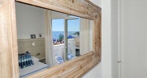 A room with a wooden-framed window, a bed with blue and white bedding, a bedside table, and a view of the sea and buildings. - Studio with sea views in the center of Calella (Calella de Palafrugell)