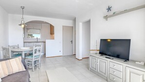 A modern living room with a flat-screen TV, a dining area with chairs, and a kitchen with a window. - Sea View Apartment in Calella, Ideal for Couples (Calella de Palafrugell)