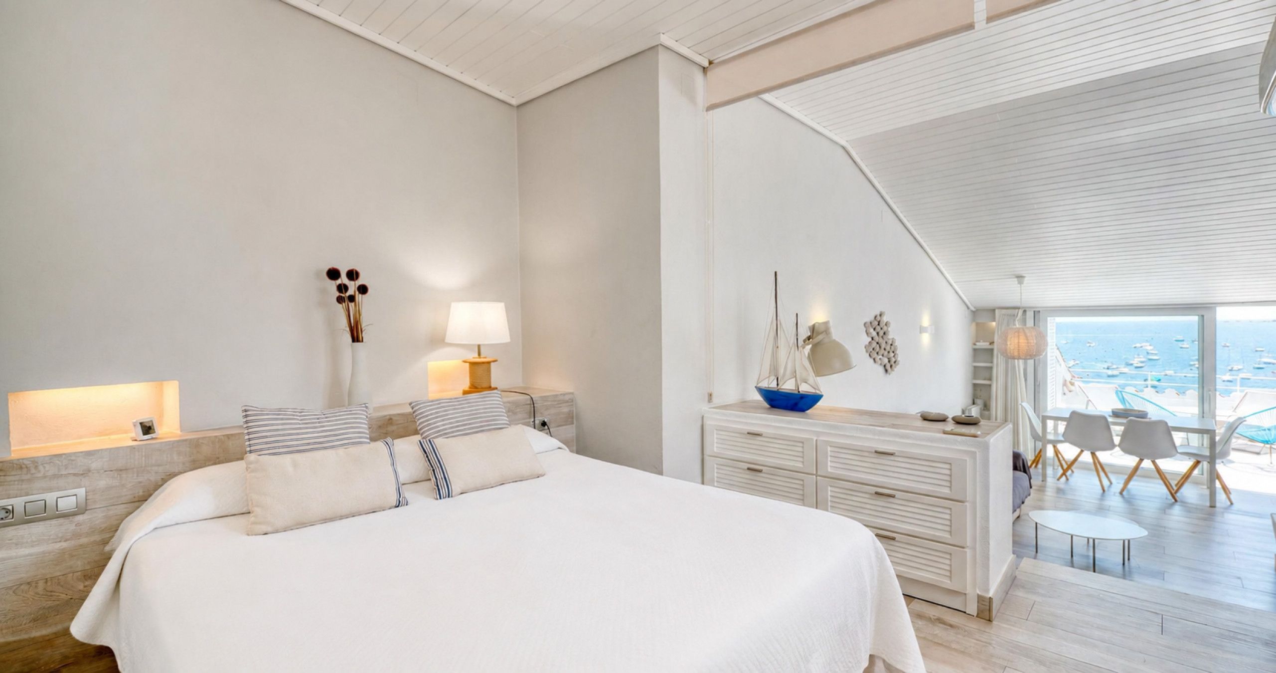 Une chambre moderne avec un grand lit, des lampes de chevet, une commode et une vue sur la mer depuis un balcon.