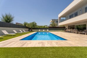 A modern outdoor swimming pool area with a wooden deck, lounge chairs, and a contemporary building in the background.