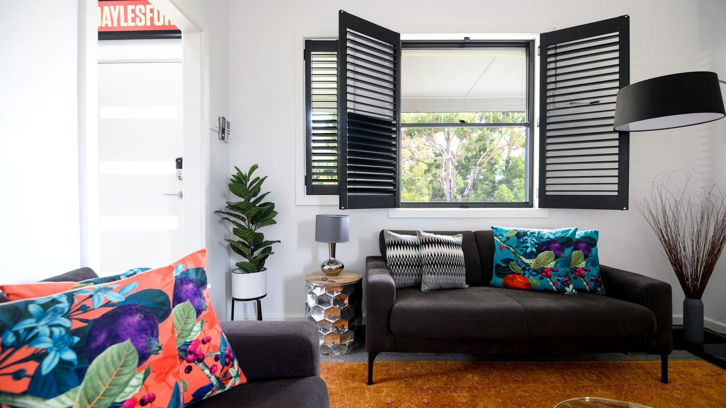 En moderne stue med sofa, to sofaborde, en potteplante og et vindue med skodder.