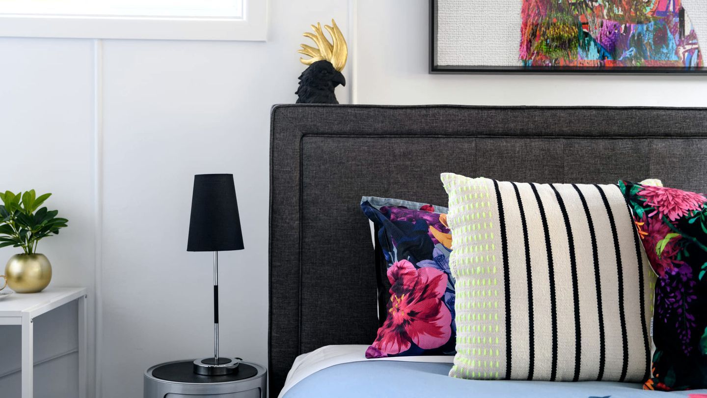A neatly made bed with a grey headboard, a black floor lamp, and a small round table with a plant beside it.