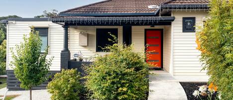 Eenlaags huis met een rode deur, een klein portaaltje en een tuin vol planten en bloemen.
