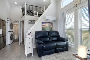 A modern living room with a black leather sofa, a staircase, an air conditioning unit, and a view of the outdoors through large windows. - Stylish Tiny Home with Fire Pit & Stargazing Deck Near Fredericksburg, TX (Fredericksburg)