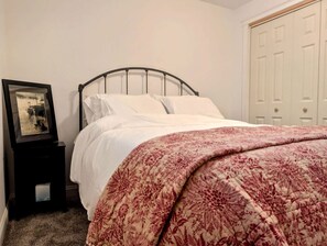 A neatly made bed with white bedding and a floral-patterned blanket, a small bedside table with a framed picture, and a white door in the background. - Coastal Beach House with Fireplace & Pool Table in Franklin, Maine (Franklin)