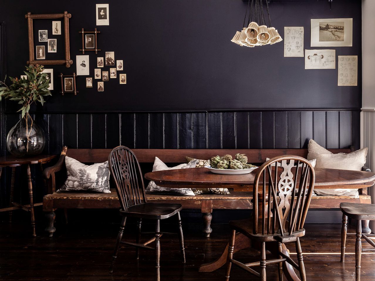A dark-themed dining area with a wooden bench, chairs, a table with a plate of artichokes, and a wall adorned with framed pictures.