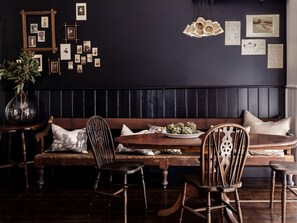 A dark-themed dining area with a wooden bench, chairs, a table with a plate of artichokes, and a wall adorned with framed pictures. - Cute Wooden Cabin with Fireplace in Daylesford, Victoria (Daylesford)