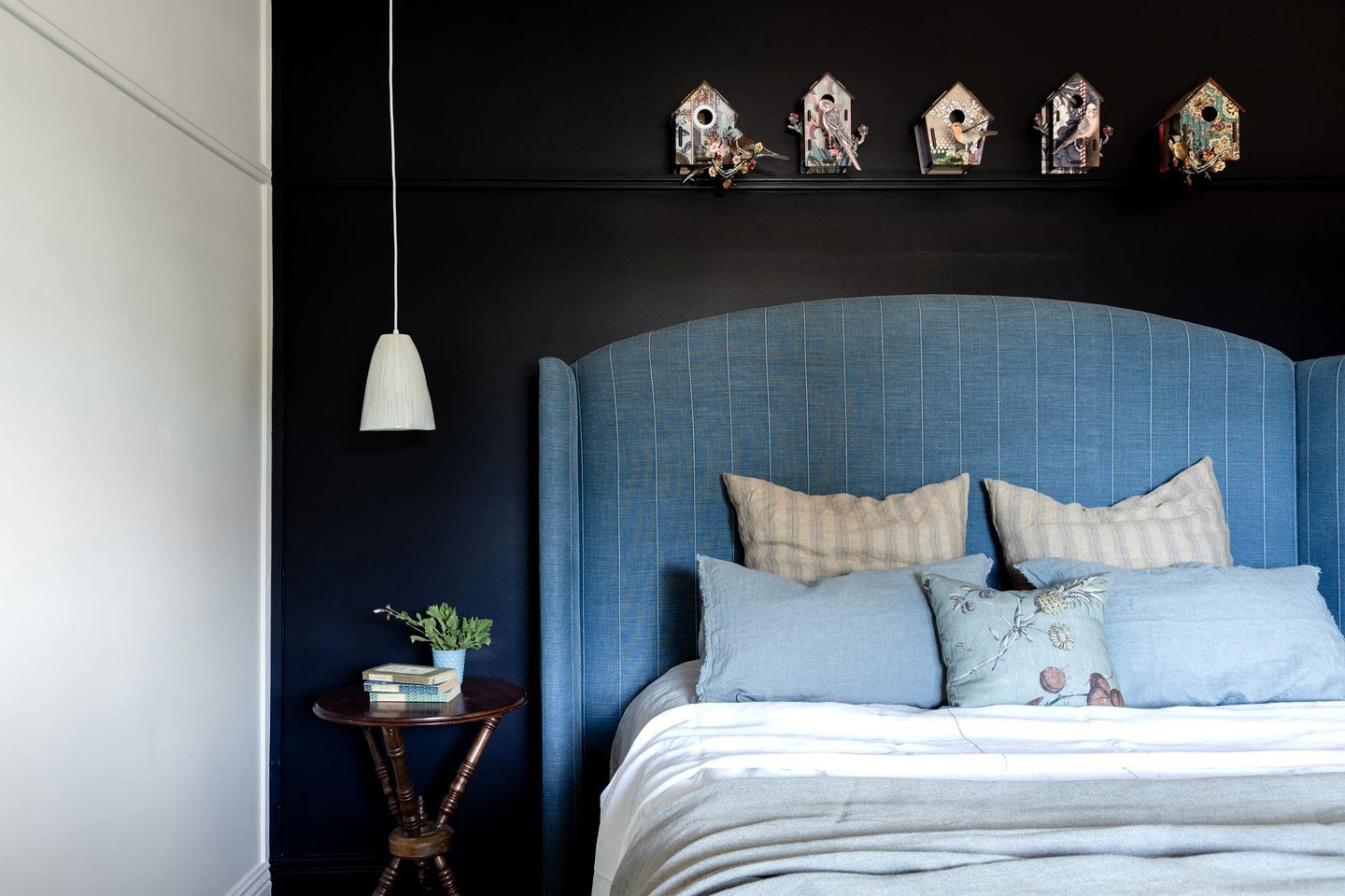 A bedroom with a blue upholstered headboard, a bedside table with a plant, and decorative birdhouses on the wall.
