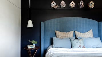 A bedroom with a blue upholstered headboard, a bedside table with a plant, and decorative birdhouses on the wall.